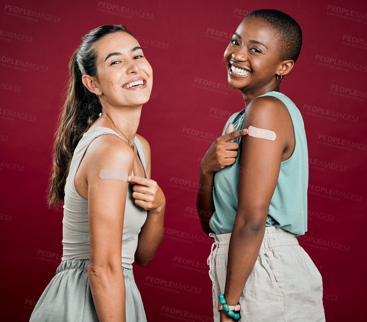 Buy stock photo Covid vaccinated African american and mixed race women showing, pointing at arm plaster. Two happy people isolated on red studio background with copyspace. Black woman and hispanic with corona vaccine