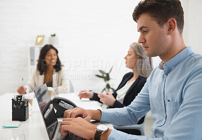 Buy stock photo Young focused businessman typing on his laptop in an office at work. Mature caucasian businesswoman talking to an african american colleague while a male coworker works on his laptop