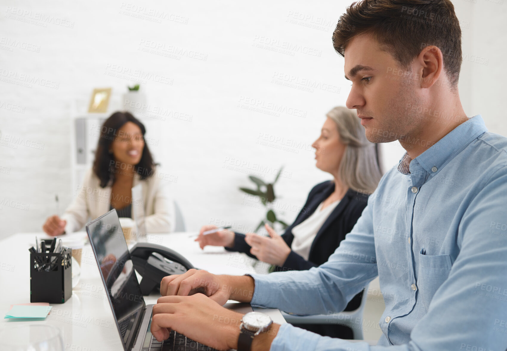 Buy stock photo Young focused businessman typing on his laptop in an office at work. Mature caucasian businesswoman talking to an african american colleague while a male coworker works on his laptop