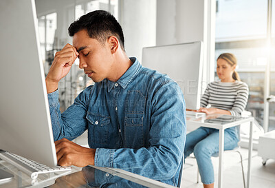 Young stressed asian businessman suffering from a headache while working on a desktop computer at work. One unhappy chinese male businessperson suffering from anxiety while working on a computer at a desk in an office