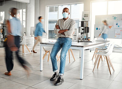 Young African american businessman standing with his arms crossed and wearing a mask at work. One confident male manager wearing a mask protecting from a virus and standing in a busy office at work