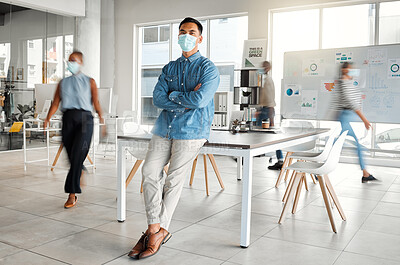 Young asian businessman standing with his arms crossed and wearing a mask at work. One confident chinese male boss wearing a mask protecting from a virus and standing in a busy office at work