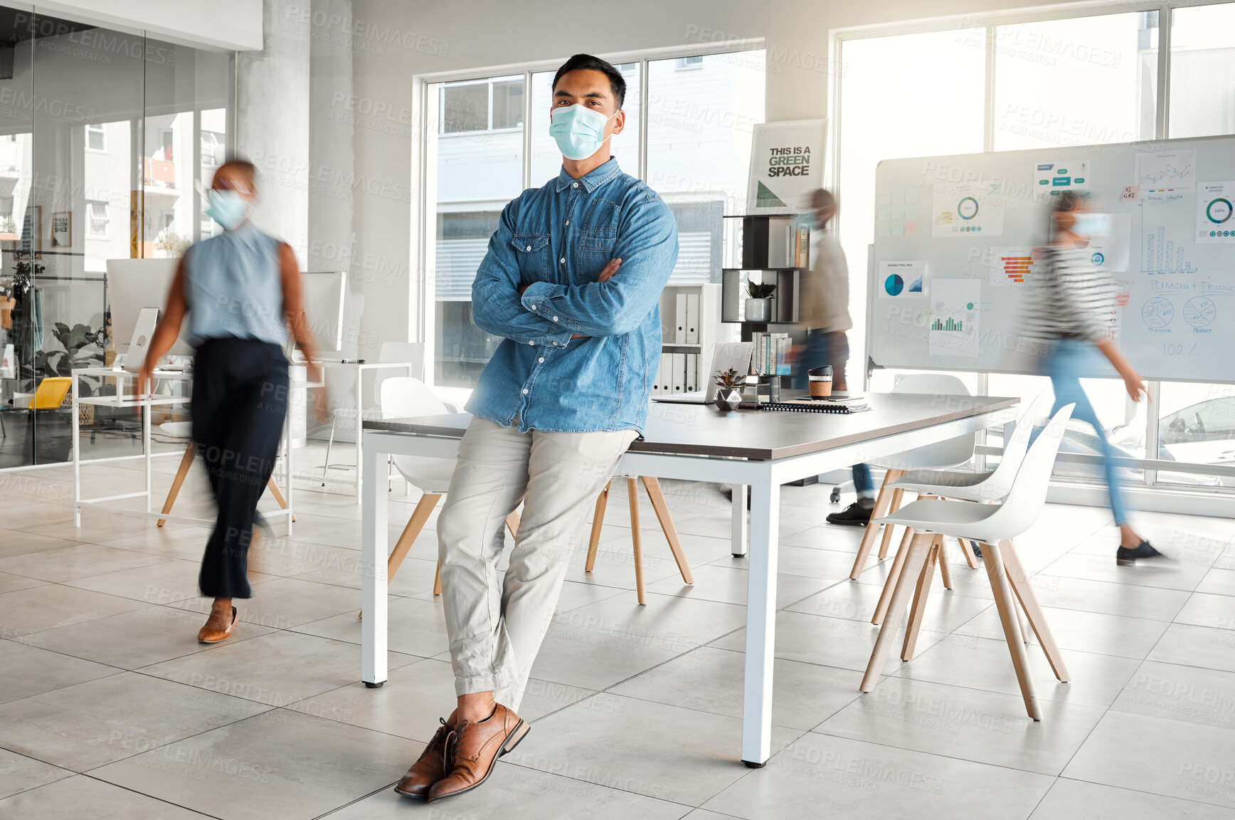 Buy stock photo Young asian businessman standing with his arms crossed and wearing a mask at work. One confident chinese male boss wearing a mask protecting from a virus and standing in a busy office at work