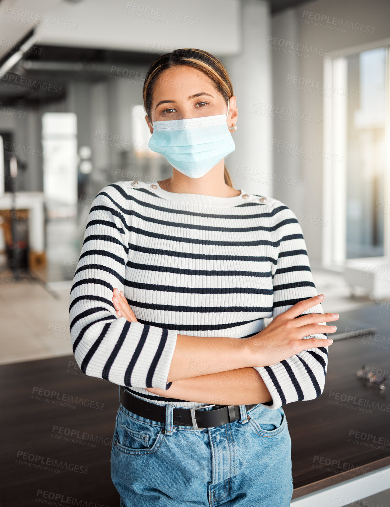 Buy stock photo Young mixed race businesswoman standing with her arms crossed and wearing a mask at work. One hispanic female boss wearing a mask protecting from a virus and standing at work