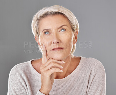One beautiful caucasian mature woman isolated against a grey studio background, posing with a hand on her cheek and thinking about making a decision. Ageing woman touching her face while wishing