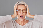 Portrait of one beautiful caucasian mature woman isolated against a grey studio background and expressing  shock while wearing glasses. Senior woman making facial expressions and looking surprised