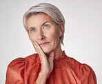 One beautiful caucasian mature woman isolated against a grey studio background, posing with a hand on her cheek and thinking about making a decision. Ageing woman touching her face while wishing