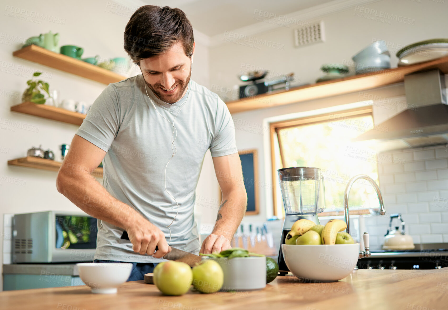Buy stock photo One fit young caucasian man cutting ingredients with knife to make healthy green detox smoothie while wearing earphones in kitchen at home. Guy having fresh fruit juice to cleanse and provide energy for training. Wholesome drink with vitamins and nutrients