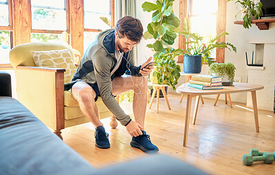 Buy stock photo One fit young caucasian man getting dressed for exercise workout while listening to music with earphones on a cellphone at home. Guy sitting on a chair putting on sports shoe sneakers while getting ready to go to gym for training