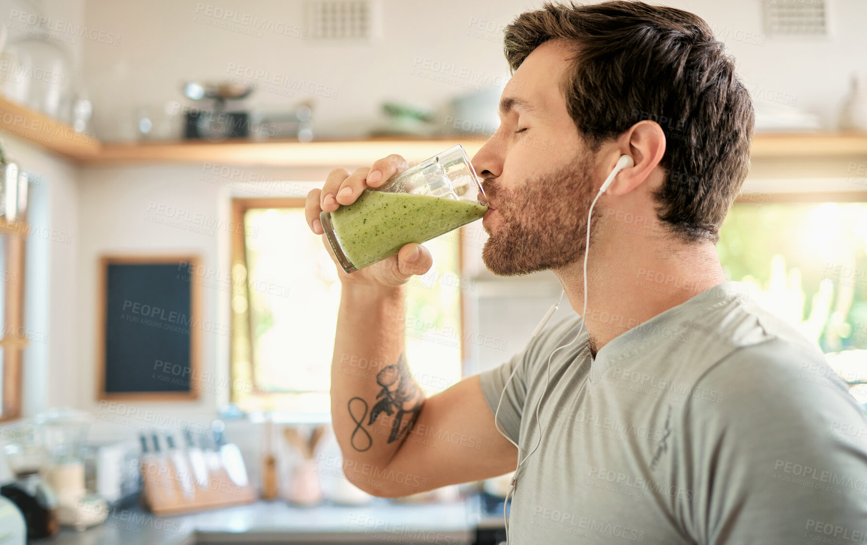 Buy stock photo One fit young caucasian man drinking a glass of healthy green detox smoothie while wearing earphones in a kitchen at home. Guy having fresh fruit juice to cleanse and provide energy for training. Wholesome drink with vitamins and nutrients