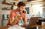 Young content caucasian businessman on a call using a phone while working on a laptop at home alone. Happy male businessperson smiling and talking on a cellphone while working in the kitchen at home