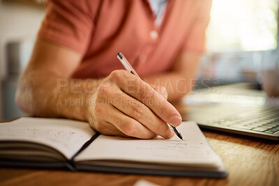 Man writing in a diary while working from home alone. One businessperson working on a laptop and writing in a notebook with a pen sitting at a table in the morning at home