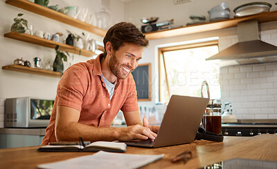 Young cheerful caucasian businessman working on a laptop at home alone. Happy male businessperson smiling and typing an email on a computer while working in the kitchen at home