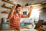 Young cheerful caucasian businessman cheering with joy while working on a laptop at home alone. One joyful male businessperson smiling while working in the kitchen at home