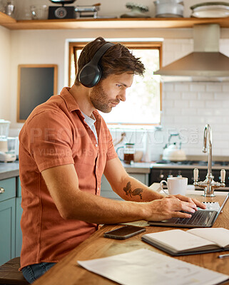 Young serious caucasian businessman wearing headphones and listening to music while using a laptop at home alone. One male businessperson typing on a laptop while working in the kitchen at home