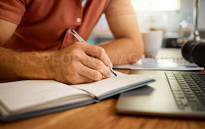 Man writing in a diary while working from home alone. One businessperson working on a laptop and writing in a notebook sitting at a table in the morning at home