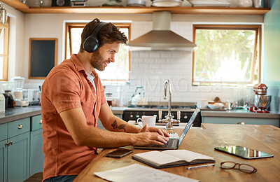 Young happy caucasian businessman wearing headphones and listening to music while using a laptop at home alone. One content male businessperson typing on a laptop while working in the kitchen at home