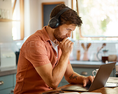 Young serious caucasian businessman wearing headphones and listening to music while using a laptop at home alone. One focused male businessperson typing on a laptop while working in the kitchen at home