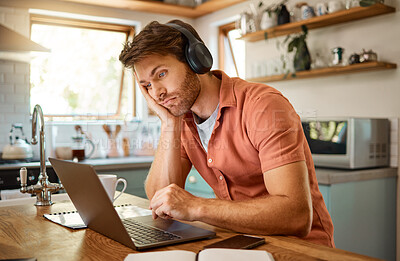 Young serious caucasian businessman wearing headphones and listening to music while working on a laptop from home alone. One stressed businessman looking unhappy while using a laptop. Person looking upset and worried