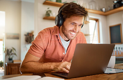 Young cheerful caucasian businessman wearing headphones and listening to music while using a laptop at home alone. One content male businessperson typing on a laptop while working in the kitchen at home