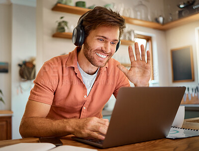 Young happy caucasian businessman wearing headphones and waving his hand on a virtual meeting on a laptop at home alone. One joyful male businessperson smiling while working in the kitchen at home
