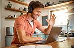 Young happy caucasian businessman wearing headphones and greeting on a virtual meeting on a laptop at home alone. One joyful male businessperson smiling while working in the kitchen at home