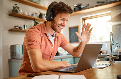 Young happy caucasian businessman wearing headphones and greeting on a virtual meeting on a laptop at home alone. One joyful male businessperson smiling while working in the kitchen at home