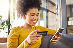 Young happy mixed race woman using a credit card and phone alone at home. Content hispanic female with a curly afro making an online purchase with a debit card and cellphone while sitting on the couch at home
