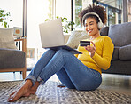 Young happy mixed race woman using a credit card and laptop while wearing headphones and listening to music alone at home. One hispanic female with a curly afro making an online purchase with a debit card and laptop while sitting on the floor at home