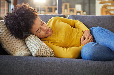 Young unhappy mixed race woman holding her stomach and suffering from stomach pain on the couch at home. One sick hispanic woman suffering from cramps resting on the sofa