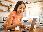 Young happy mixed race woman using a credit card and laptop while relaxing alone at home. One content hispanic female making an online payment and typing on a computer while sitting in her kitchen