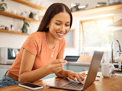 Young happy mixed race woman using a credit card and laptop while relaxing alone at home. One content hispanic female making an online payment and typing on a computer while sitting in her kitchen