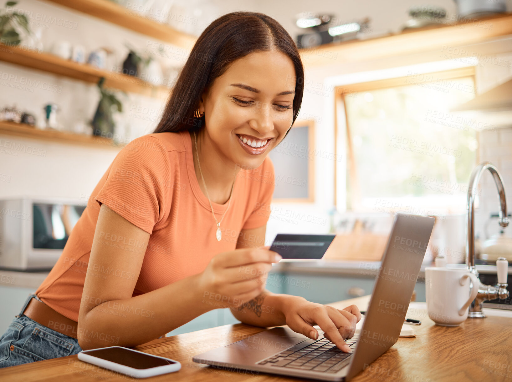 Buy stock photo Young happy mixed race woman using a credit card and laptop while relaxing alone at home. One content hispanic female making an online payment and typing on a computer while sitting in her kitchen