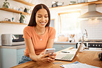 Young beautiful happy mixed race businesswoman holding and using a phone while working from home. One content hispanic woman using social media on her cellphone while using a laptop sitting at a kitchen counter