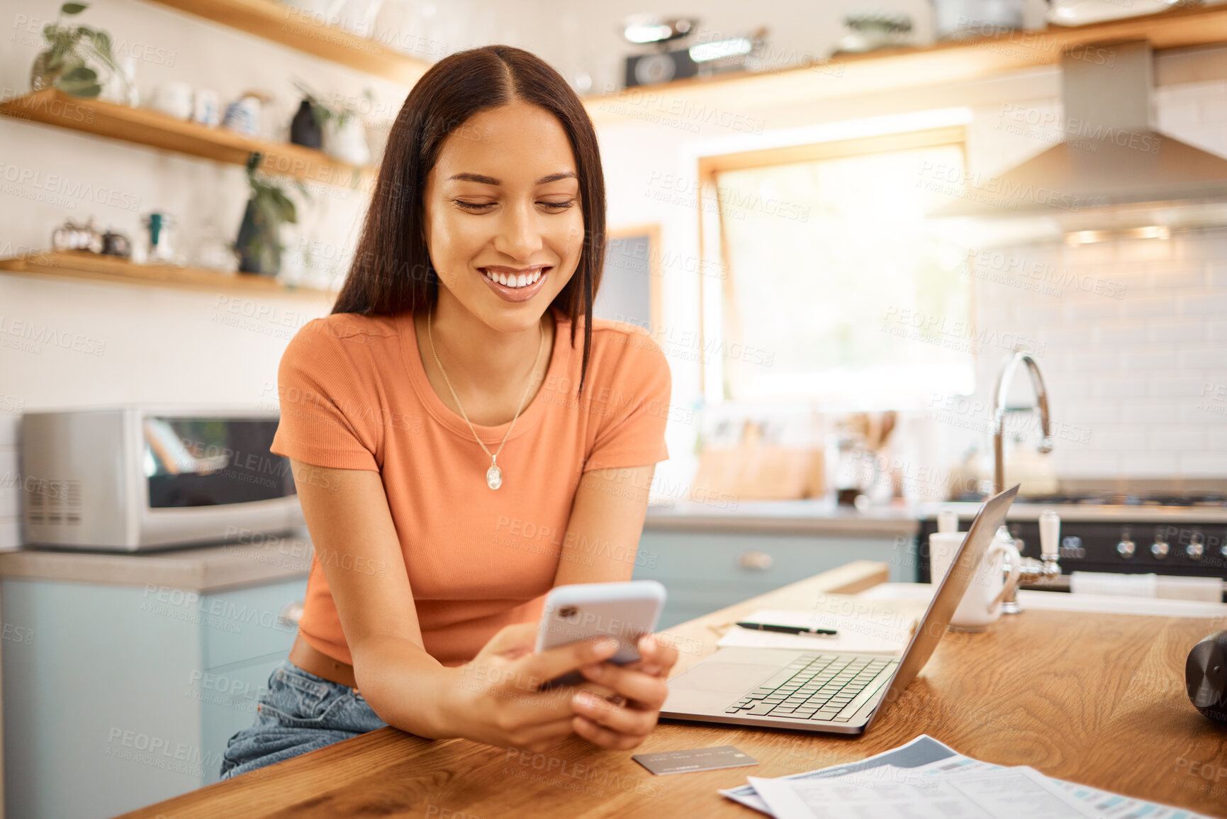 Buy stock photo Young beautiful happy mixed race businesswoman holding and using a phone while working from home. One content hispanic woman using social media on her cellphone while using a laptop sitting at a kitchen counter