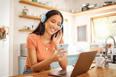 Young beautiful cheerful mixed race businesswoman holding and using a phone while wearing headphones and listening to music working from home. One joyful hispanic woman using social media on her cellphone and enjoying a music break while working on a lapt
