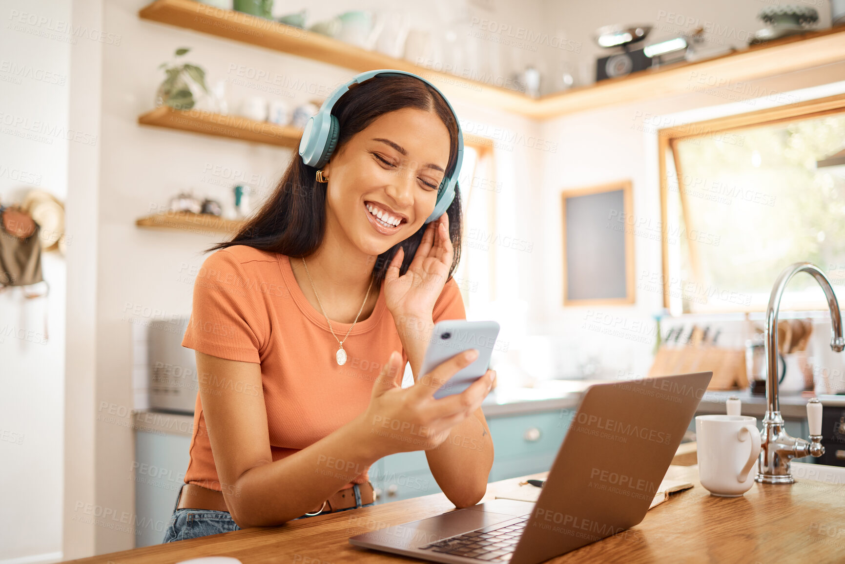 Buy stock photo Young beautiful cheerful mixed race businesswoman holding and using a phone while wearing headphones and listening to music working from home. One joyful hispanic woman using social media on her cellphone and enjoying a music break while working on a laptop