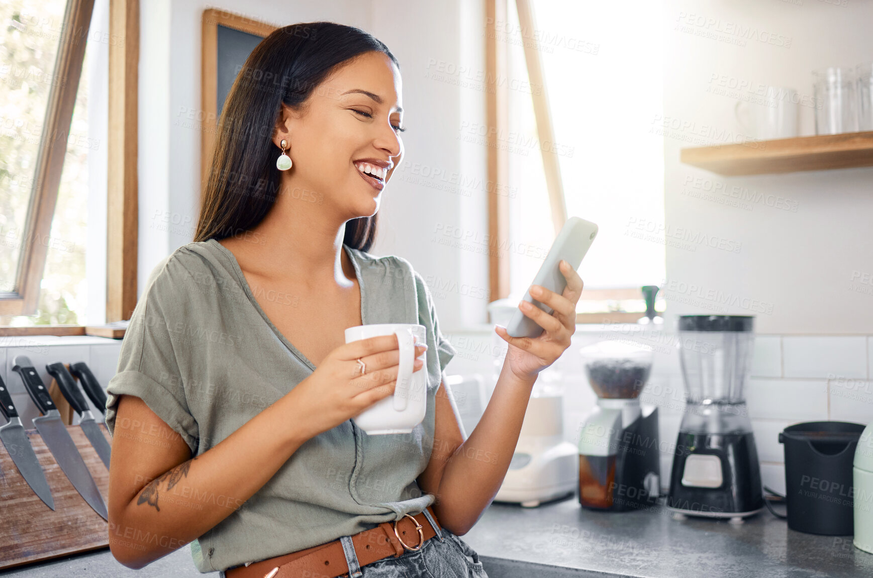 Buy stock photo Young mixed race hispanic woman using a phone while drinking coffee in the morning at home. Happy woman using social media on her phone standing in the kitchen at home and laughing at funny video
