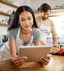 Close up of beautiful young woman browsing on digital tablet while her boyfriend cooks in the background. Young hispanic female searching for recipe to follow online while cooking at home with her husband