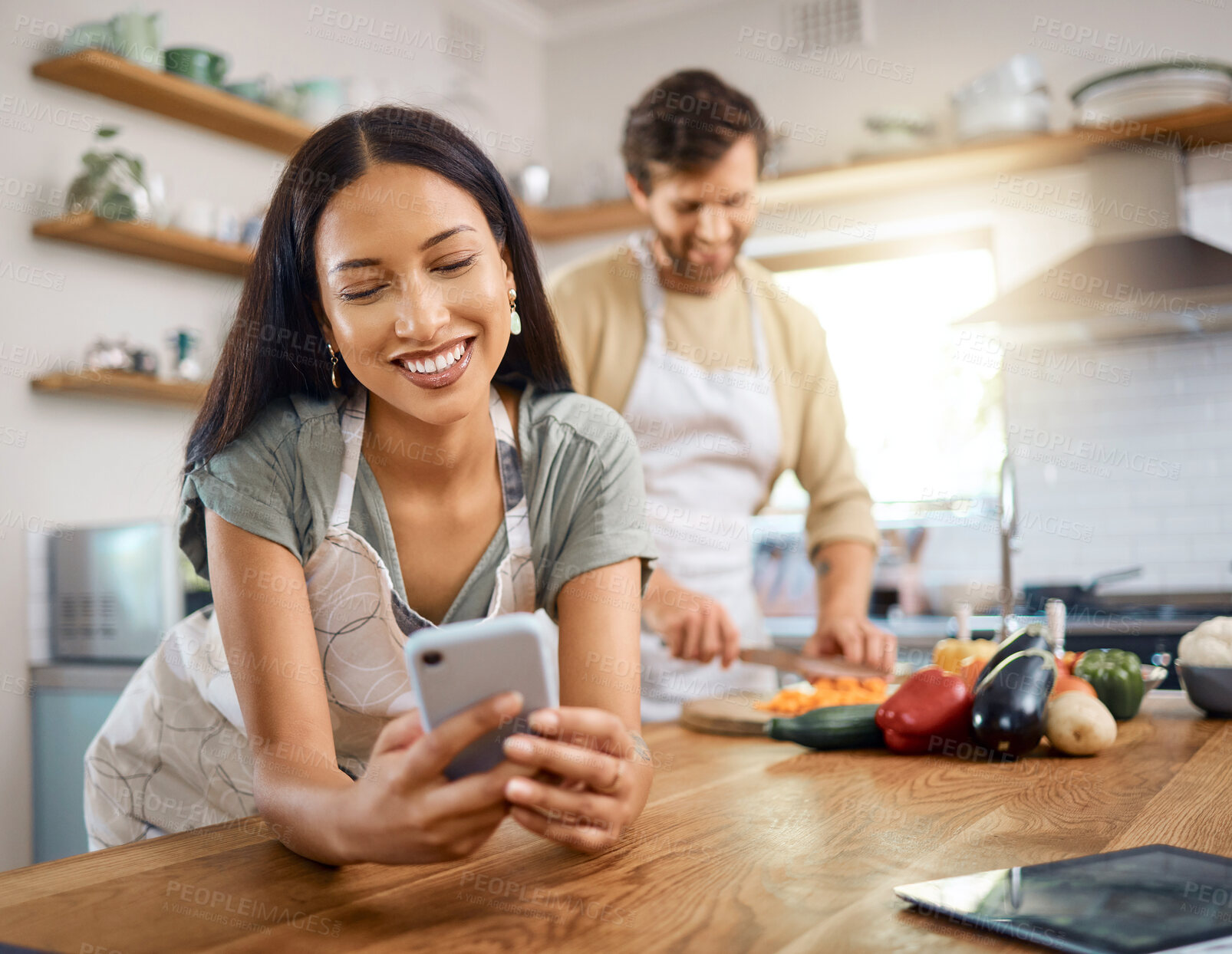 Buy stock photo Happy young woman browsing social media on smartphone while her boyfriend cooks in the background. Young hispanic female searching for recipe on internet or sending text message while cooking at home with her husband