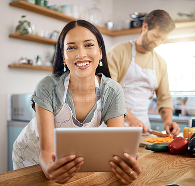 Buy stock photo Portrait of beautiful young woman browsing on digital tablet while her boyfriend cooks in the background. Young hispanic female searching for recipe to follow online while cooking at home with her husband