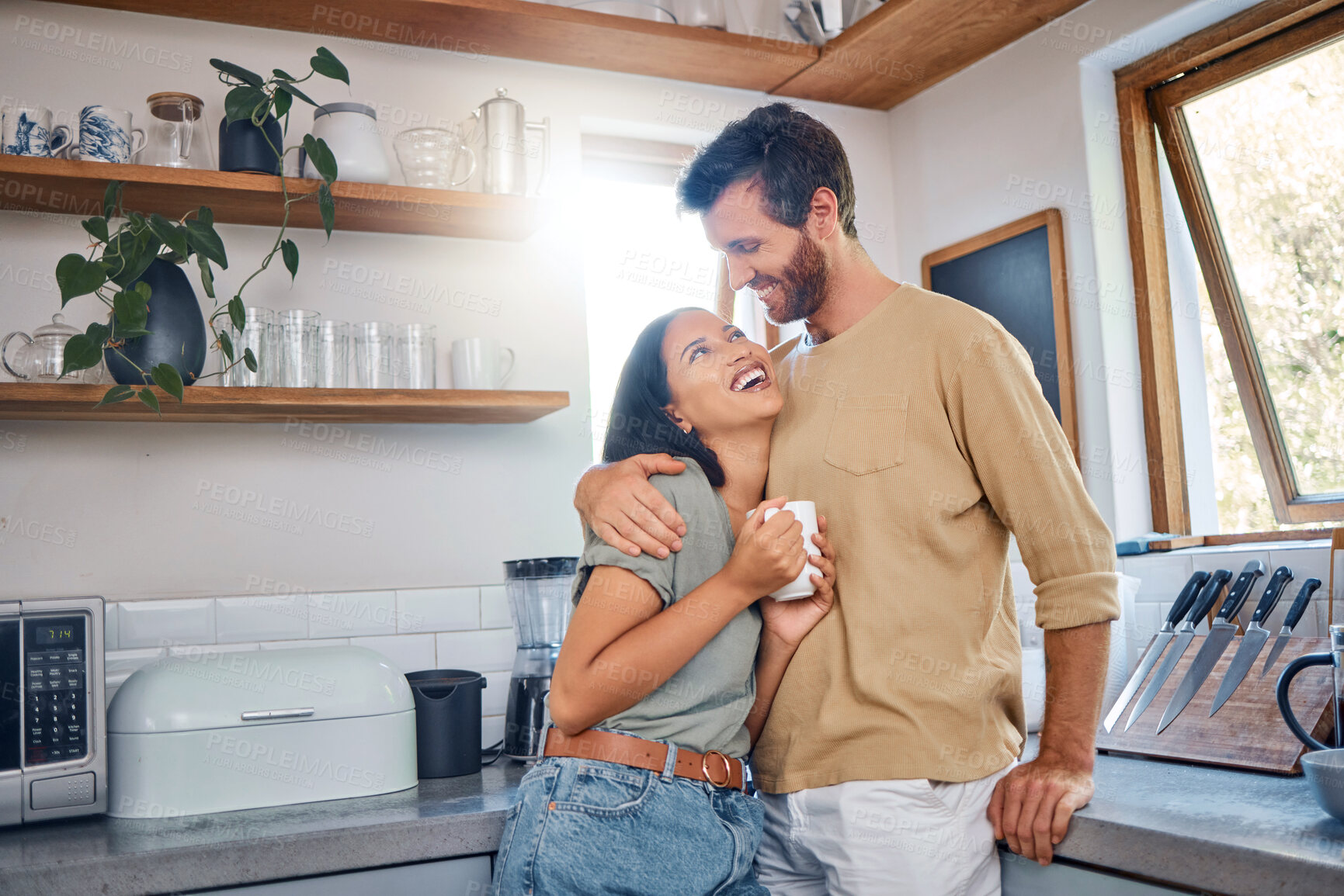 Buy stock photo Happy young interracial couple spending time together at home. Young woman enjoying her morning coffee while boyfriend puts his arms around her. Young couple showing love and affection while enjoying morning leisure time