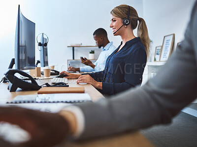 Happy young caucasian call centre telemarketing agent talking on a headset while working on computer alongside colleagues in an office. Confident friendly female consultant operating helpdesk for customer service and sales support