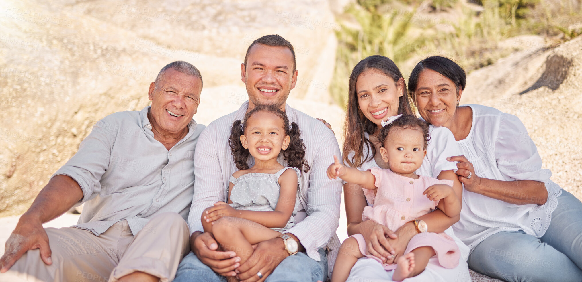 Buy stock photo Portrait of a smiling mixed race family with little girls sitting  together at the beach. Adorable little kids bonding with their parents and grandparents while having a picnic at a park or garden