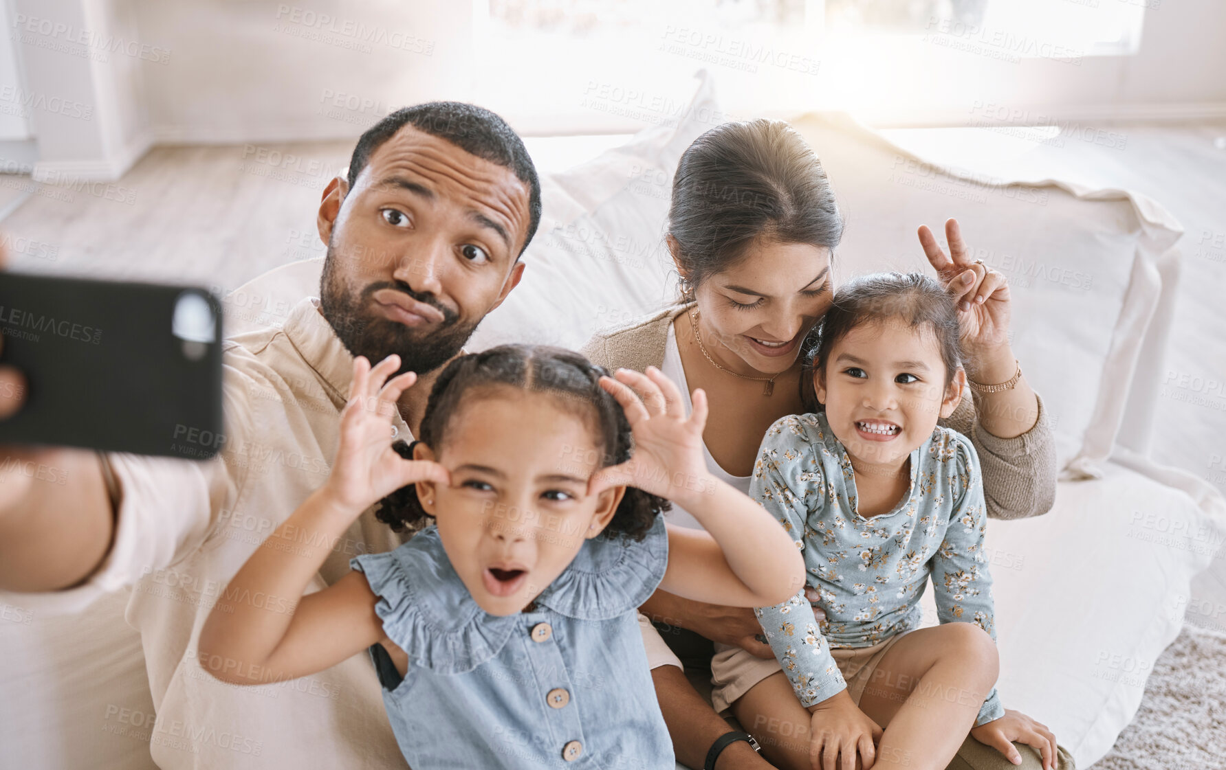 Buy stock photo Portrait of a mixed race family taking a selfie and making funny faces while sitting on the sofa at home. Hispanic man using a smartphone to take a photo with his wife and two little cute daughters in the lounge