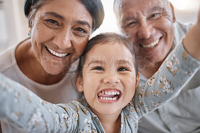 Portrait of smiling mixed race grandparents and granddaughter taking a selfie in the lounge at home. Hispanic senior man and woman taking photos and bonding with their cute little granddaughter at home