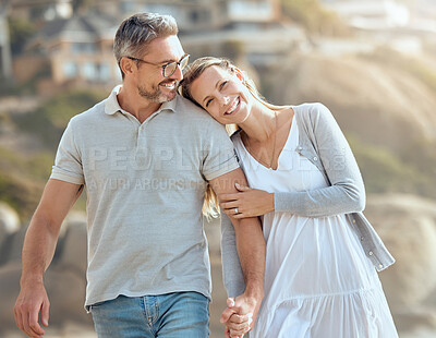 Happy and loving mature caucasian couple enjoying a romantic walk at the beach together on a sunny day. Cheerful affectionate wife resting on husband\'s shoulder while holding hands and bonding on vacation outdoors