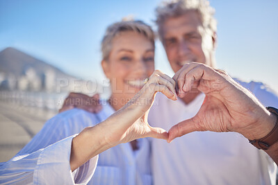 Buy stock photo A happy mature caucasian couple enjoying fresh air on vacation at the beach while bonding. Smiling retired couple hugging and showing a heart shape with their fingers