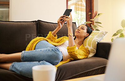 Woman using her smartphone lying on a sofa, relaxing and resting in a bright living room. A young hispanic female texting on her cellphone using modern technology at home. Isolated during lockdown