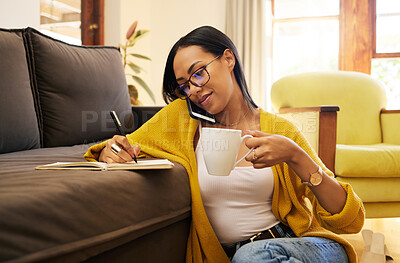 Hispanic woman writing notes sitting on the floor against a sofa drinking coffee in a bright living room. A serious focused young female working remotely during a work meeting on a phone call at home